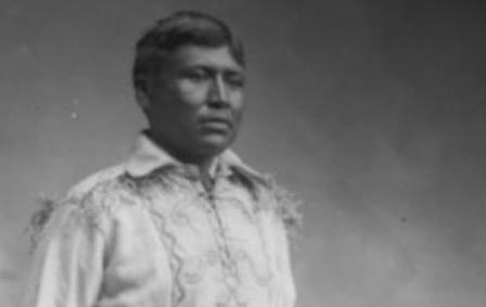 A photo of Chief Jim Boss standing in traditional clothes with one hand on the back of the chair