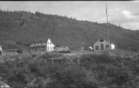 Archival black and white photo of buildings on a grassy area with a hilly mountain behind them.