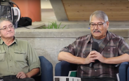 Two older men sit in chairs as one speaks into a microphone