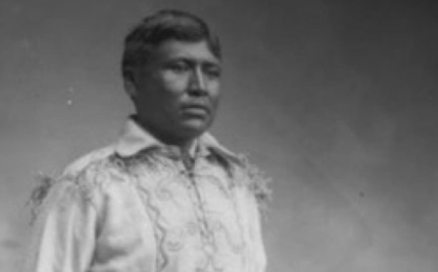 A photo of Chief Jim Boss standing in traditional clothes with one hand on the back of the chair