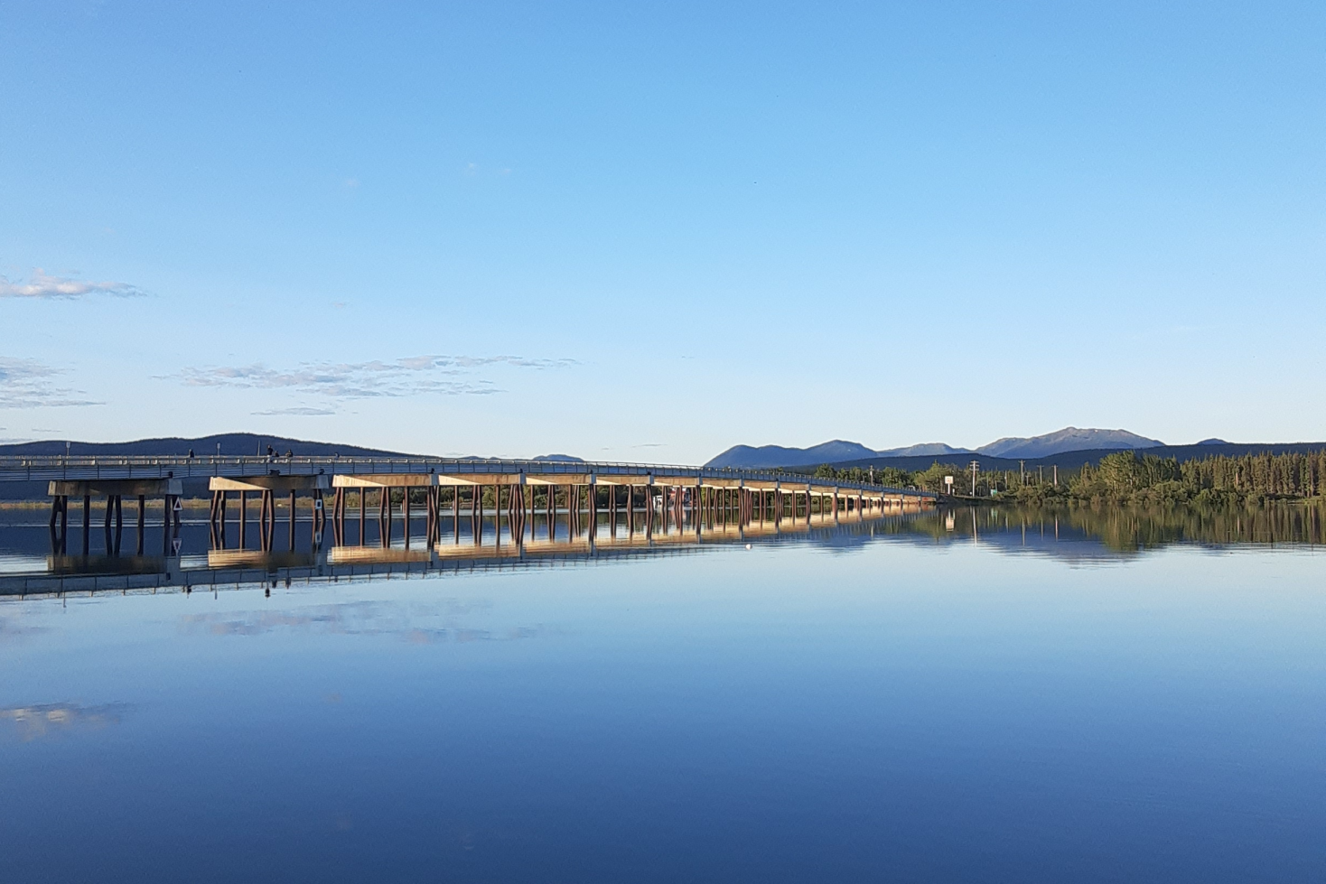 A bridge is against the horizon in the middle of the image with the sky above and water of the Tagish River in the foreground, trees in background.